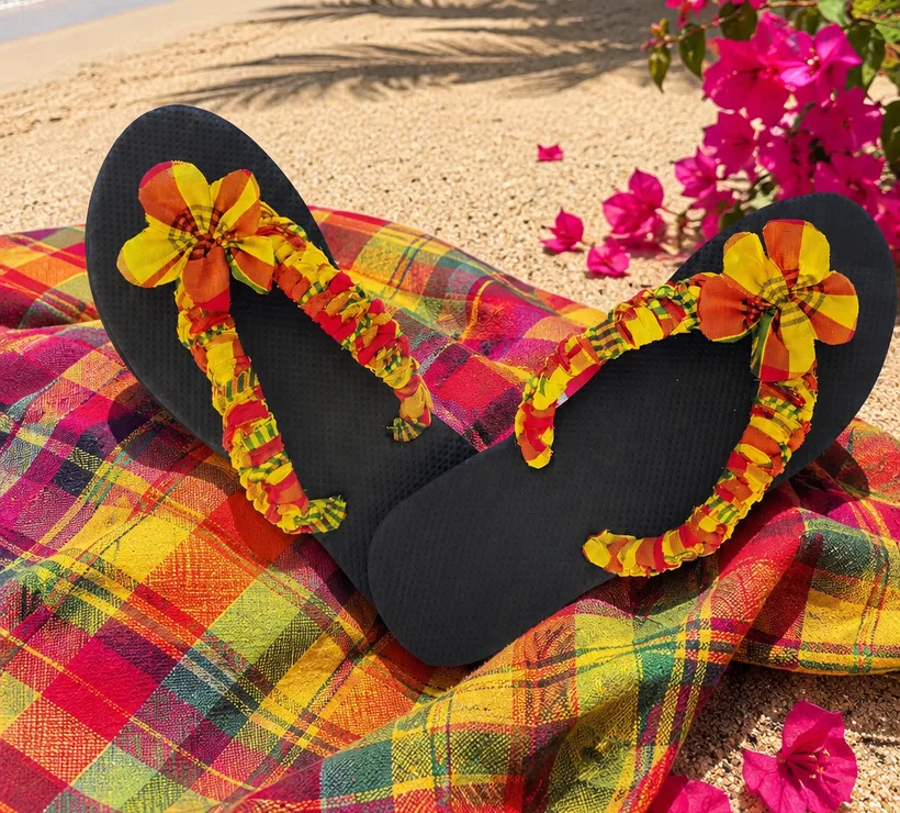 Tongs madras noires faites main posées sur un tissu madras, sur le sable d'une plage de Guadeloupe